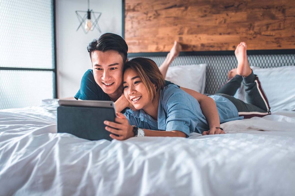 How well do you know your partner? Image of couple on bed looking at tablet together and smiling.